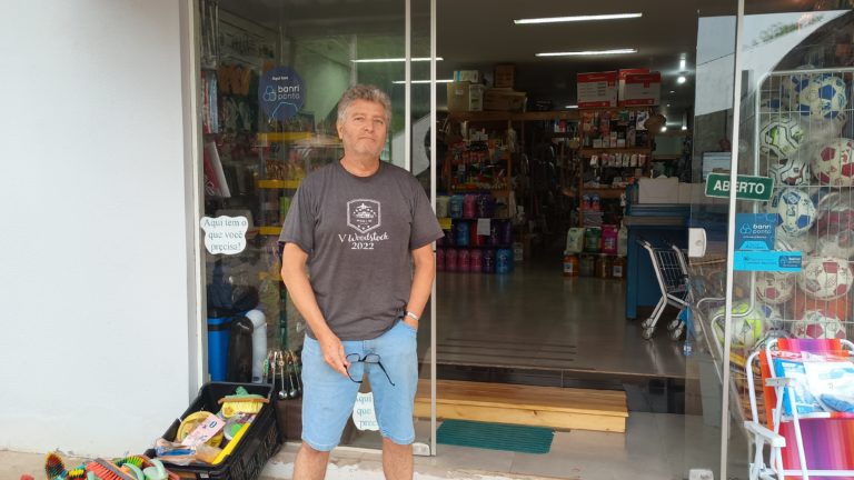 Foto colorida de Leonardo Bastiani, em frente à fachada de sua loja no centro de Muçum. Ele é um homem branco, com cabelo grisalho, usa uma camiseta cinza e bermuda jeans