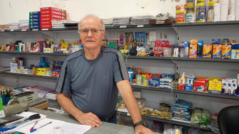 Foto colorida de Vilmar Zilío, em seu bazar e papelaria no centro de Muçum. Ele é um homem branco, idoso, com cabelo branco, usa óculos e camiseta cinza com detalhes pretos