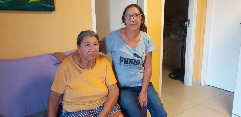 Foto colorida de Neli e Nadia dos Santos Silva. Neli é uma mulher negra idosa, com cabelo grisalho e usa uma blusa laranja. Nadia é uma mulher negra, usa óculos e tem cabelo preto. Usa uma blusa cinza.