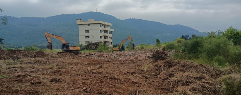 Foto colorida que mostra área com cases que foram demolidas e máquinas fazendo a remoção de escombros. Apenas um prédio aparece de pé