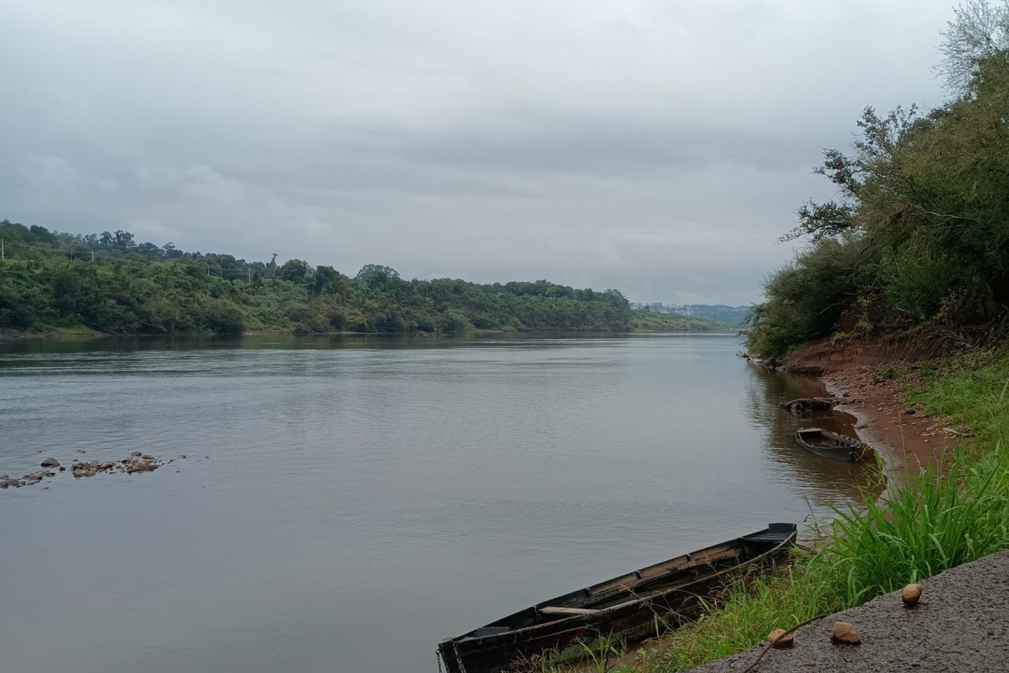 Foto colorida do Rio Taquari, em Arroio do Meio. Na imagem, está nublado e aparece uma canoa próxima da margem do Rio