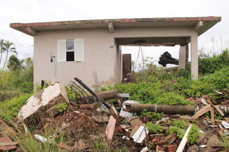 Foto colorida de casa destruída pelas enchentes, com escombros na frente e um tronco de árvore que aparece atrás de uma área aberta