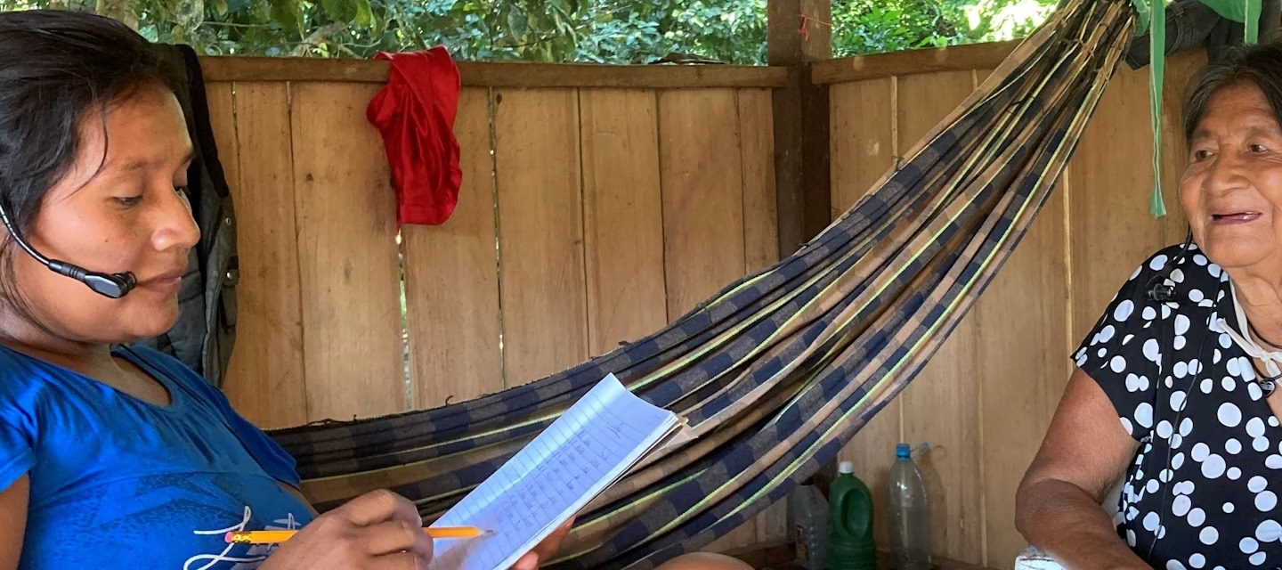 Foto colorida de Silvania Oro Eo’ Cabixi entrevista sua avó Maria Pin Wa Oro Win na aldeia São Luiz, para elaboração de um dicionário. Silvana é indígena e aparece fazendo anotações em um caderno e com um microfone no ouvido. Maria está de frente para ela, no canto direito, e sorri.