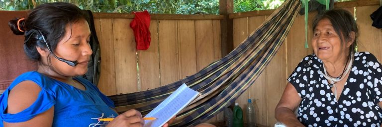 Foto colorida de Silvania Oro Eo’ Cabixi entrevista sua avó Maria Pin Wa Oro Win na aldeia São Luiz, para elaboração de um dicionário. Silvana é indígena e aparece fazendo anotações em um caderno e com um microfone no ouvido. Maria está de frente para ela, no canto direito, e sorri.