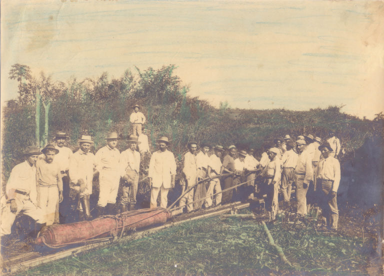 Foto colorida que mostra diversas pessoas em pé diante de tronco de madeira. Transporte de uma peça de ferro, calibre 24, retirada do Forte do Príncipe da Beira com destino ao Museu Nacional, no Rio de Janeiro, 6/07/1913. Costa Marques, Rondônia / Acervo DPHDM .