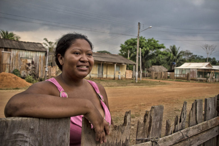Foto colorida de mulher negra escorada em cerca de madeira. Ela sorri e olha para frente. Ao fundo, casas e postes de energia. Comunidade Quilombolas do Forte Príncipe da Beira, no Vale do Guaporé, em Costa Marques, em Rondônia. (Foto: Marcela Bonfim/Amazônia Real/2018).