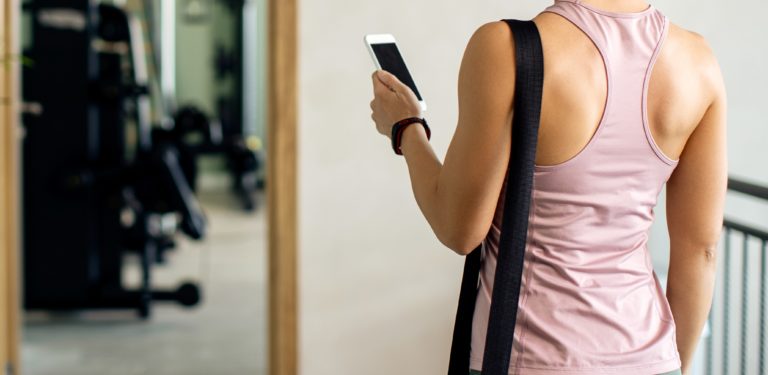 Foto colorida que mostra mulher de costas mexendo no celular. Ela está usando uma blusa rosa e, no espelho, aparecem equipamentos de academia. Ilustração de busca pelo shape