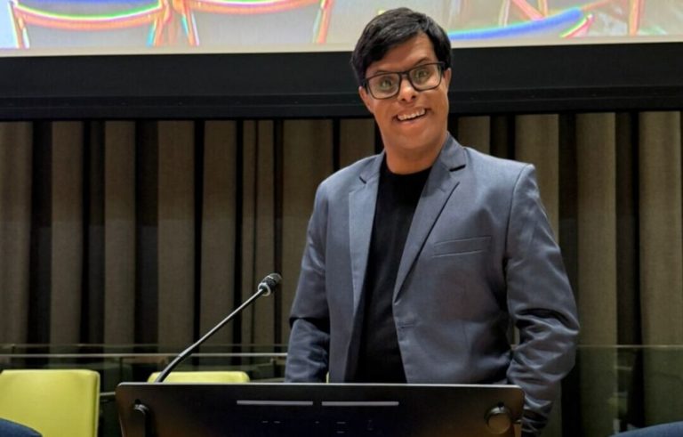 Foto colorida de Gabriel Camargos. Ele usa um terno cinza, com camiseta preta e está diante de um púlpito