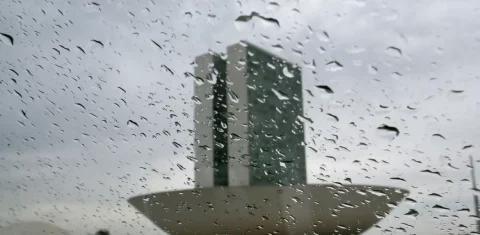Foto colorida que mostra lente ou vidro com gotas de chuva e no fundo, o prédio do Congresso Nacional. Mais ao fundo, nuvens carregadas