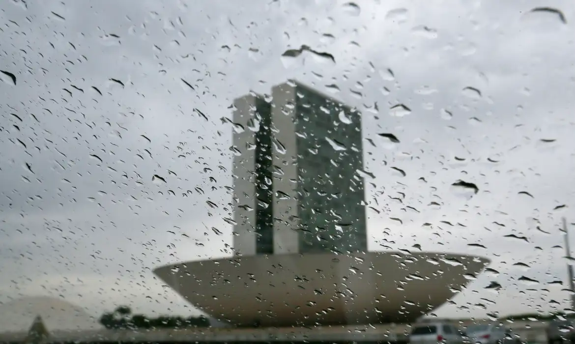 Foto colorida que mostra lente ou vidro com gotas de chuva e no fundo, o prédio do Congresso Nacional. Mais ao fundo, nuvens carregadas