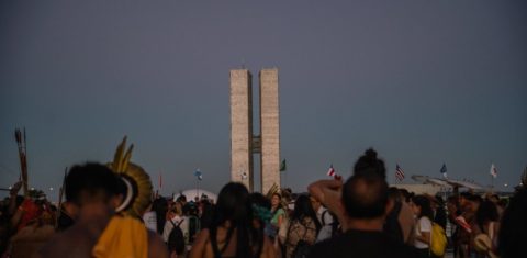 Foto colorida de indígenas de costas durante o Acampamento Terra Livre de 2023, em Brasília. Ao fundo, prédio do Congresso