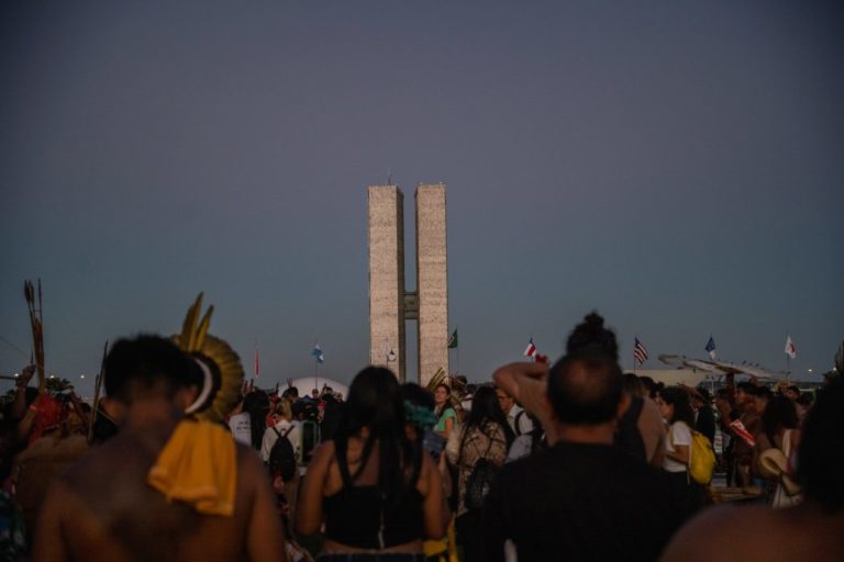 Foto colorida de indígenas de costas durante o Acampamento Terra Livre de 2023, em Brasília. Ao fundo, prédio do Congresso