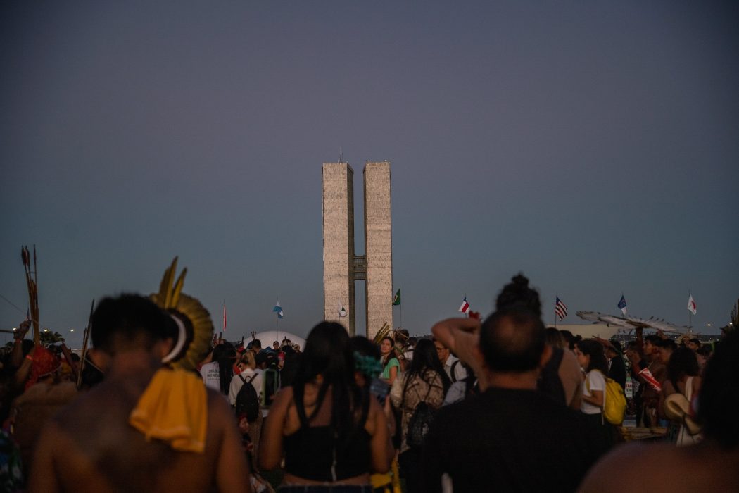 Foto colorida de indígenas de costas durante o Acampamento Terra Livre de 2023, em Brasília. Ao fundo, prédio do Congresso