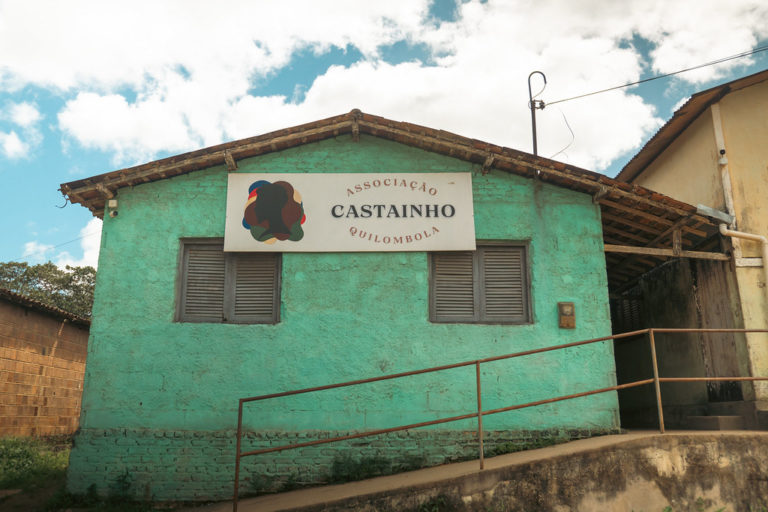 Foto colorida da fachada da Associação do Quilombo Castainho. Na imagem, aparece o prédio na cor verde, com a placa com o nome da associação