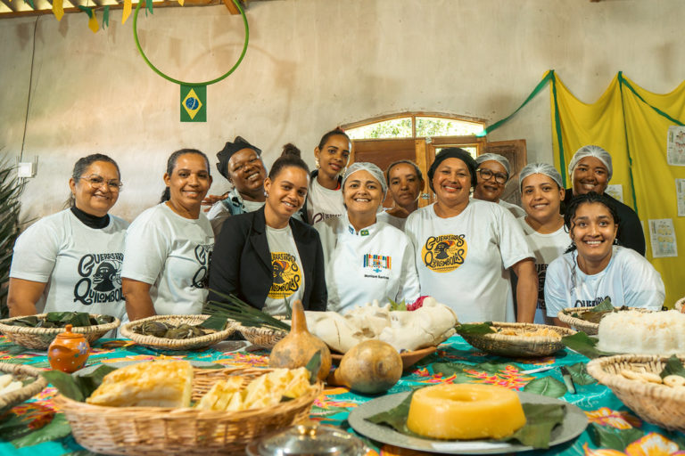 Foto colorida com grupo de mulheres da Associação de Mulheres Guerreiras Quilombolas do Castainho. Elas são quilombolas, negras, usam uma camiseta branca da associação e estão atrás de uma mesa com várias comidas feitas a partir de mandioca