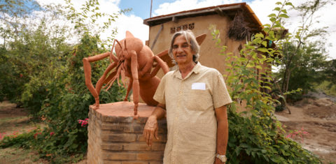 Foto colorida de Lula do Mel. Ele é um homem branco, com cabelo grisalho na altura do ombro. Ele está ao lado de uma escultura de uma abelha, no Solar das Abelhas, em Caruaru.