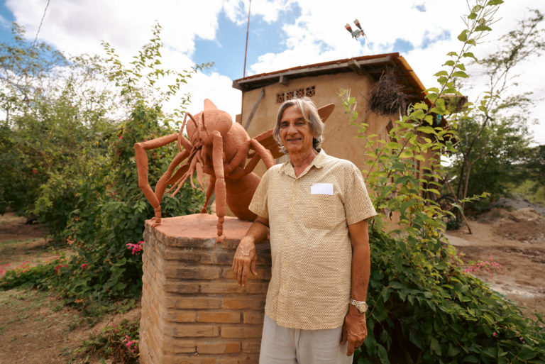 Foto colorida de Lula do Mel. Ele é um homem branco, com cabelo grisalho na altura do ombro. Ele está ao lado de uma escultura de uma abelha, no Solar das Abelhas, em Caruaru.