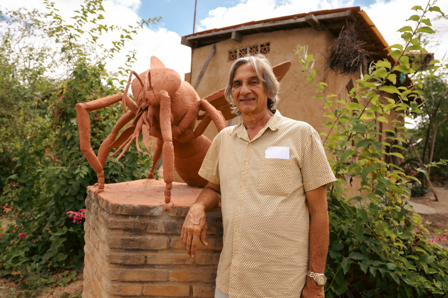 Foto colorida de Lula do Mel. Ele é um homem branco, com cabelo grisalho na altura do ombro. Ele está ao lado de uma escultura de uma abelha, no Solar das Abelhas, em Caruaru.