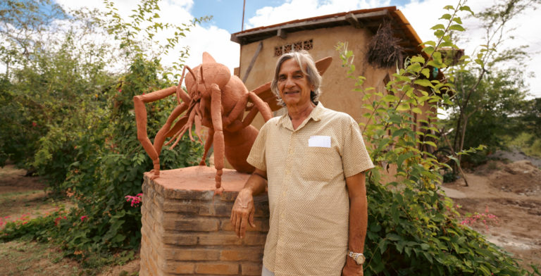 Foto colorida de Lula do Mel. Ele é um homem branco, com cabelo grisalho na altura do ombro. Ele está ao lado de uma escultura de uma abelha, no Solar das Abelhas, em Caruaru.