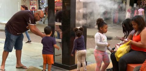 Foto colorida de pessoas em torno de refúgio climático em Belo Horizonte. Na imagem, nuvens de nebulização aparecem em volta de crianças e adultos