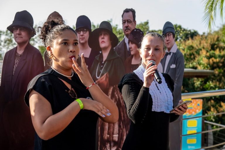 Ao lado de intérprete e à frente de painel reproduzindo a foto de Marie Curie, a professora Camila Silveira fala no evento: encontro para celebrar a existência, a resistência e a competência profissional das pesquisadoras (Foto: Filipe Dutra)