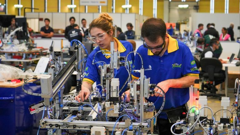 Foto colorida que mostra duas pessoas, uma mulher e um homem, ambos brancos e com camisetas azuis, em frente de equipamento industrial