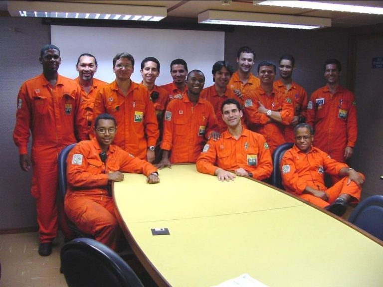 Foto colorida de grupo de trabalhadores da Petrobras. Eles aparecem em uma sala, uniformizados com roupa laranja. Alguns estão sentados e outros em pé