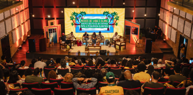 Foto colorida do painel: Modos de vida e clima, que abriu os debates da segunda edição da semana do clima na Caatinga, no Cais do Sertão, em Recife. Na imagem, aparecem os painelistas no centro e o público de costas, assistindo o evento.