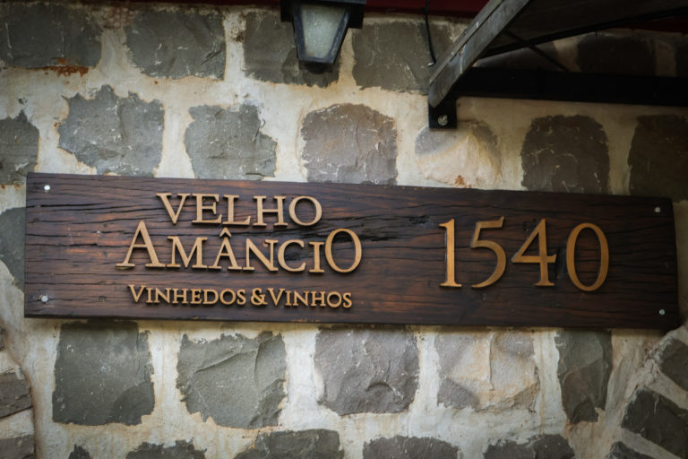 Foto colorida da placa de madeira da vinícola Velho Amâncio. Abaixo do nome, está escrito vinhedo e vinhos e, ao lado, o número 1540
