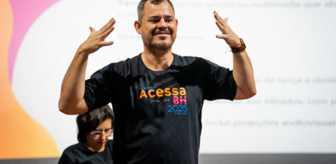 Foto colorida de Ademar durante a edição de 2025 do Festival Acessa BH, em que atua como consultor há quatro anos. Ele é um homem branco, com barba e cabelo curto, grisalhos. Ele veste uma camiseta preta, onde está escrito Acessa BH e está com os dois braços levantados, na altura da cabeça