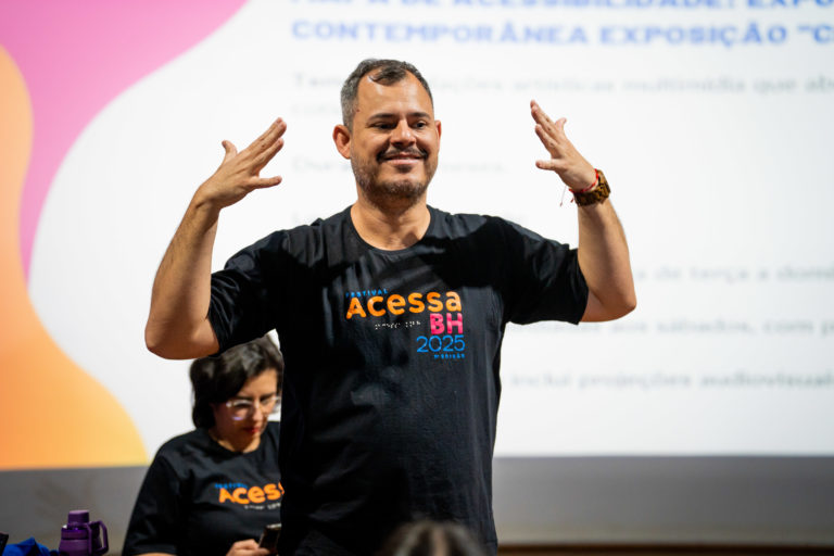 Foto colorida de Ademar durante a edição de 2025 do Festival Acessa BH, em que atua como consultor há quatro anos. Ele é um homem branco, com barba e cabelo curto, grisalhos. Ele veste uma camiseta preta, onde está escrito Acessa BH e está com os dois braços levantados, na altura da cabeça