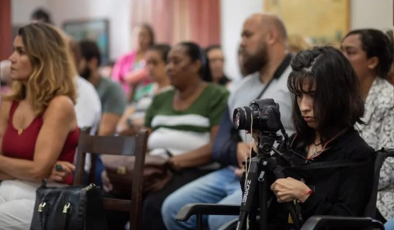 Foto colorida de Cintia Soares, usando equipamento de tripé adaptado. Ela aparece em primeiro plano, no canto direito da imagem. Ela é uma mulher branca, com cabelo preto e está segurando o tripé, sentada. Ao fundo, aparecem diversas pessoas desfocadas