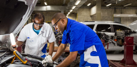 Foto colorida de pessoa mexendo em peças de motor de carro. É um homem negro que usa um jaleco azul, com um S branco. Ao lado dele, aparece um homem mais velho, usando óculos e camisa branca