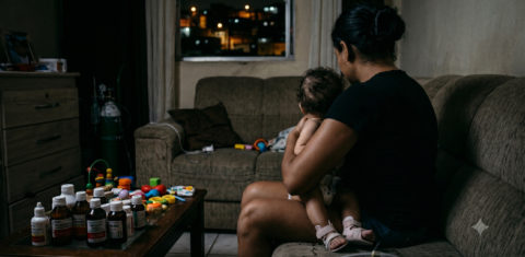 Foto colorida gerada por IA que mostra uma mãe segurando uma criança no colo. Ela está de costas e olha para um sofá, cheio de remédios, na sua frente aparece uma mesa, também cheia de medicamentos