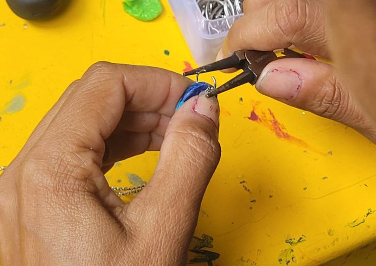 Foto colorida de um close nas mãos de uma pessoa confeccionando uma ecojoia, com mesa amarela no fundo