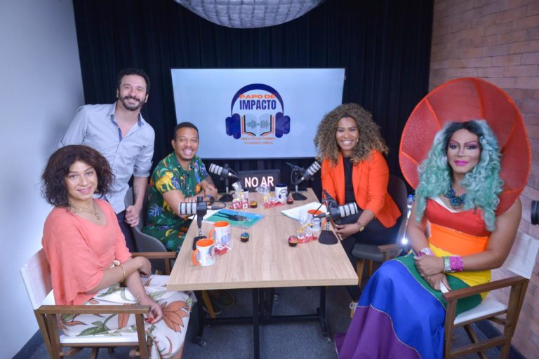 Foto colorida de Pertuzzatty durante gravação de podcast. Ela está sentada, no canto esquerdo da imagem, usando um blusão cor salmão e tem cabelos cacheados castanhos. Sobre a mesa, há microfones e canecas. Ao fundo, está uma tela que exibe uma imagem de fones de ouvido e o texto "PAPO DE IMPACTO".