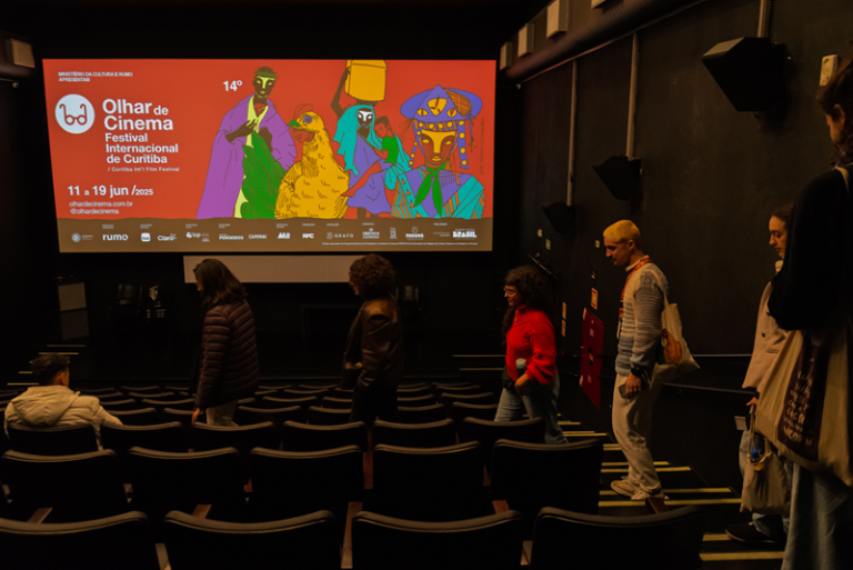 Foto colorida de pessoas entrando em sala de cinema. No telão, aparece cartaz do Olhar de Cinema – Festival Internacional de Curitiba,