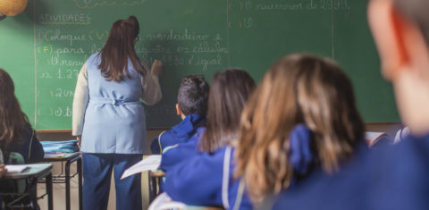Foto colorida que mostra uma professora de costas e escrevendo em um quadro. Em primeiro plano, aparecem várias crianças também de costas.