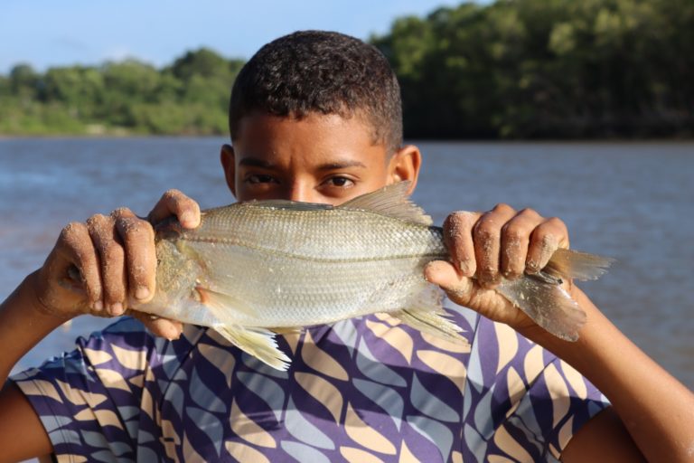 Foto colorida de criança segurando um peixe na altura da boca de modo que a parte de baixo do rosto fica encoberta. Ao fundo, rio e floresta