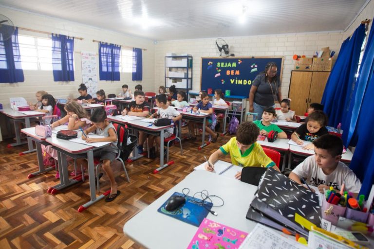 Foto colorida de sala de aula com diversas estudantes e uma professora no canto direito. Ela é uma mulher negra e está de pé, usando uma camiseta cinza
