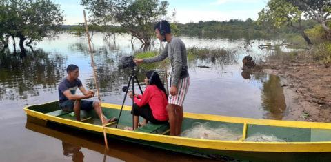 Foto colorida de pessoas em um barco. Um homem aparece em pé mexendo em câmera que está em um tripé, enquanto uma mulher entrevista outro homem, ambos sentados na canoa. Ao fundo, Rio