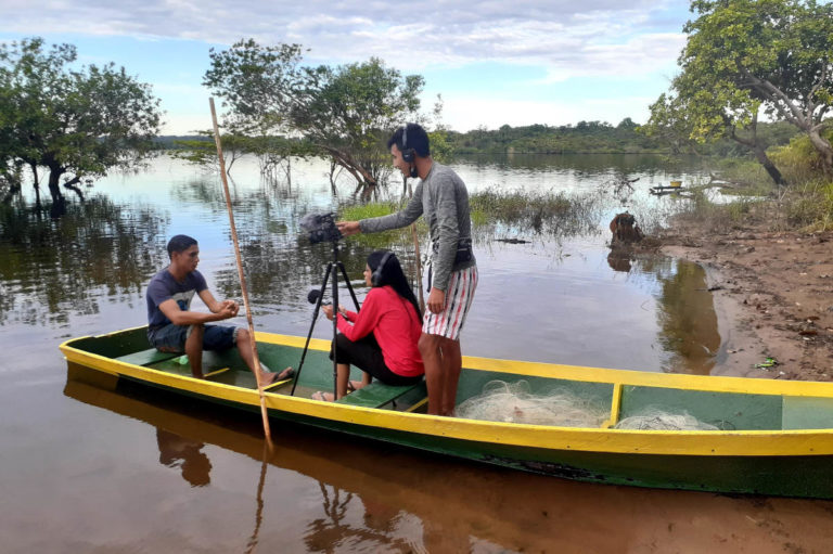 Foto colorida de pessoas em um barco. Um homem aparece em pé mexendo em câmera que está em um tripé, enquanto uma mulher entrevista outro homem, ambos sentados na canoa. Ao fundo, Rio
