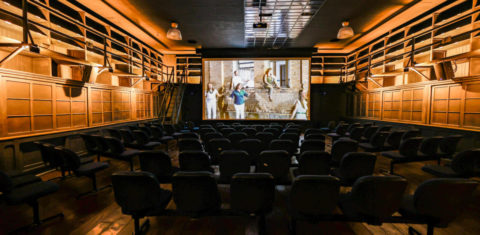 Foto colorida da sala de cinema do Espaço Expressa, em Jundiaí. O local está vazio e, ao fundo, no telão está sendo reproduzido um vídeo