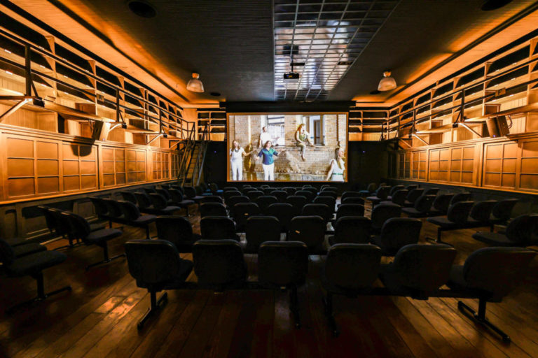 Foto colorida da sala de cinema do Espaço Expressa, em Jundiaí. O local está vazio e, ao fundo, no telão está sendo reproduzido um vídeo