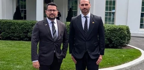 Foto colorida de Paulo Figueiredo e Eduardo Bolsonaro, de terno, em frente à Casa Branca, nos Estados Unidos. Os dois estão de pé e olham para a câmera