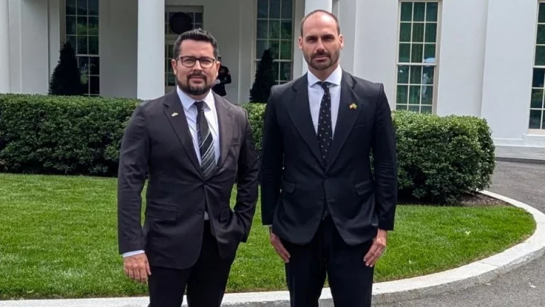 Foto colorida de Paulo Figueiredo e Eduardo Bolsonaro, de terno, em frente à Casa Branca, nos Estados Unidos. Os dois estão de pé e olham para a câmera