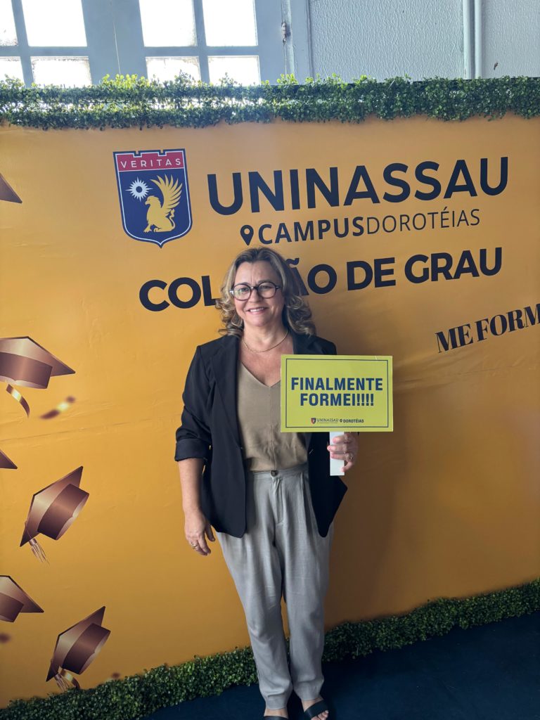 Foto colorida de Graça Alves, segurando placa que diz "finalmente formei!!!". Ela é uma mulher branca, com cabelos loiros na altura do pescoço. Ela sorri e usa óculos, vestindo um blazer preto e uma blusa bege. Ao fundo, banner com logo da Uninassau