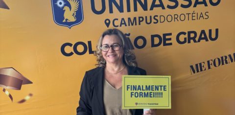 Foto colorida de Graça Alves, segurando placa que diz "finalmente formei!!!". Ela é uma mulher branca, com cabelos loiros na altura do pescoço. Ela sorri e usa óculos, vestindo um blazer preto e uma blusa bege. Ao fundo, banner com logo da Uninassau