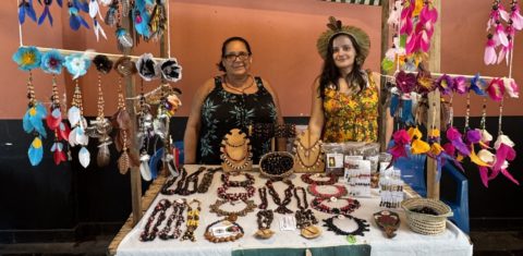 Foto colorida de Leide Calazancio ao lado da mãe, Sandra. Leide usa uma blusa amarela estampada e um cocar, Sandra uma blusa preta e óculos. Ambas são mulheres indígenas e estão diante de estande com várias biojoias e artesanato