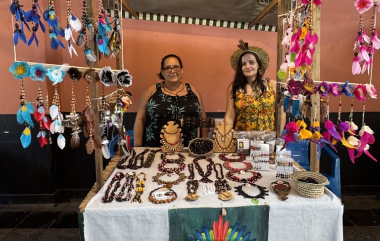 Foto colorida de Leide Calazancio ao lado da mãe, Sandra. Leide usa uma blusa amarela estampada e um cocar, Sandra uma blusa preta e óculos. Ambas são mulheres indígenas e estão diante de estande com várias biojoias e artesanato